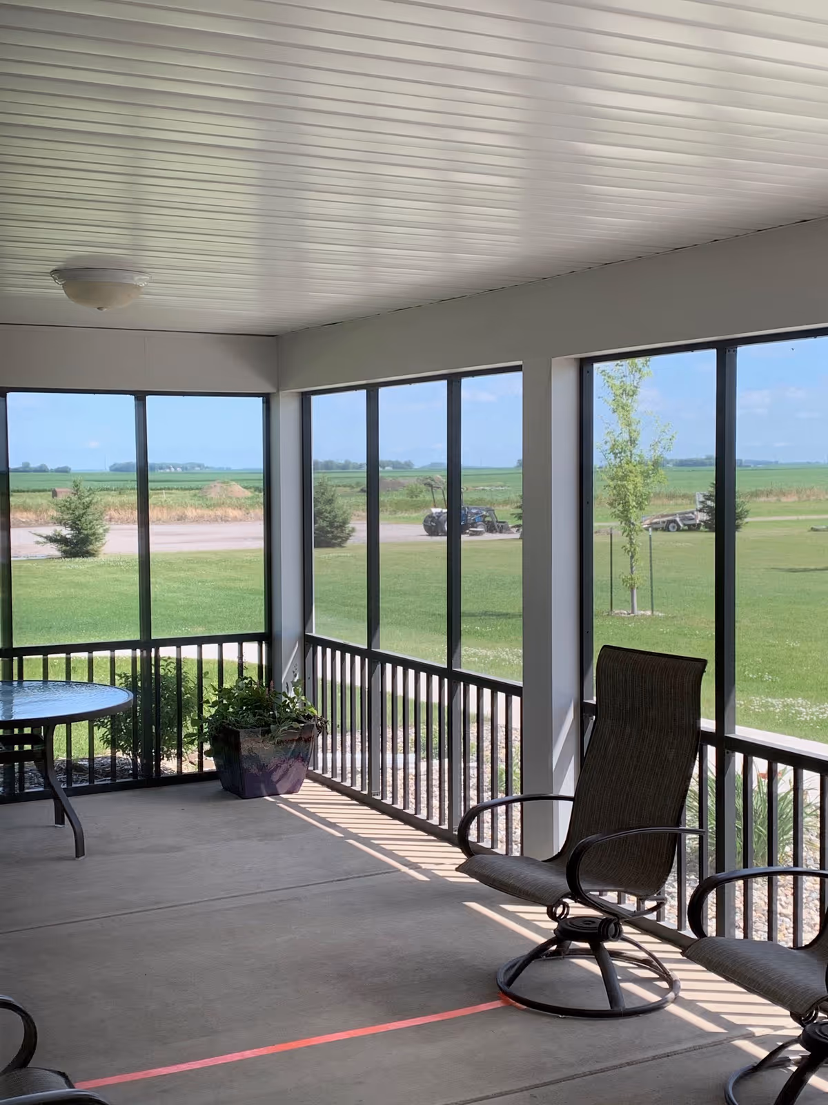 A screened-in porch area with two metal chairs and a round glass table. The porch overlooks a green lawn with a few small trees and a rural landscape in the background under a blue sky with some clouds.