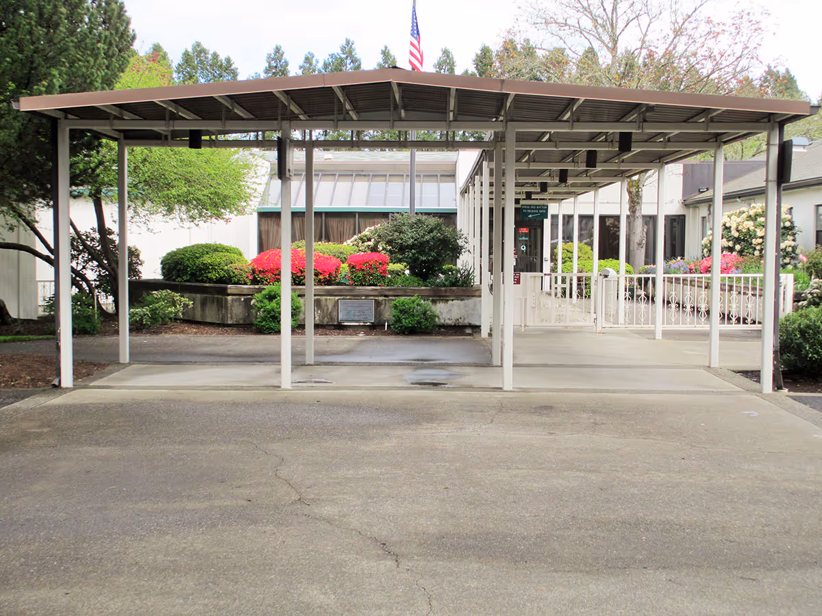 Covered entrance area with a metal roof supported by white pillars, leading to a building surrounded by well-maintained bushes and flowering plants, with an American flag visible above the roofline.