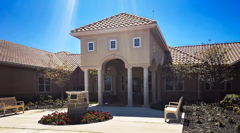 Exterior view of a senior living facility entrance with a tiled roof, beige walls, and a covered porch supported by columns. There are benches on either side of the walkway, landscaped flower beds, and small trees under a clear blue sky.