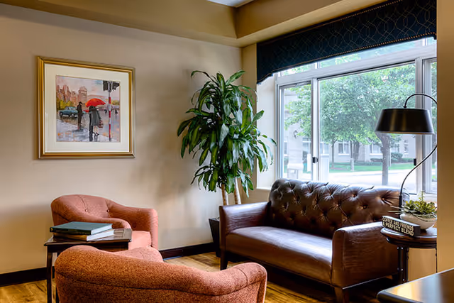 A cozy living room area with a brown leather tufted sofa, two upholstered armchairs in a reddish fabric, a side table with books and a lamp, a large green potted plant, and a framed painting on the beige wall. A large window with a dark valance lets in natural light and shows trees and a building outside.