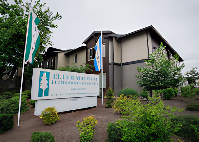 Front exterior of a two-story senior living building with a large entrance sign and flags surrounded by shrubs.