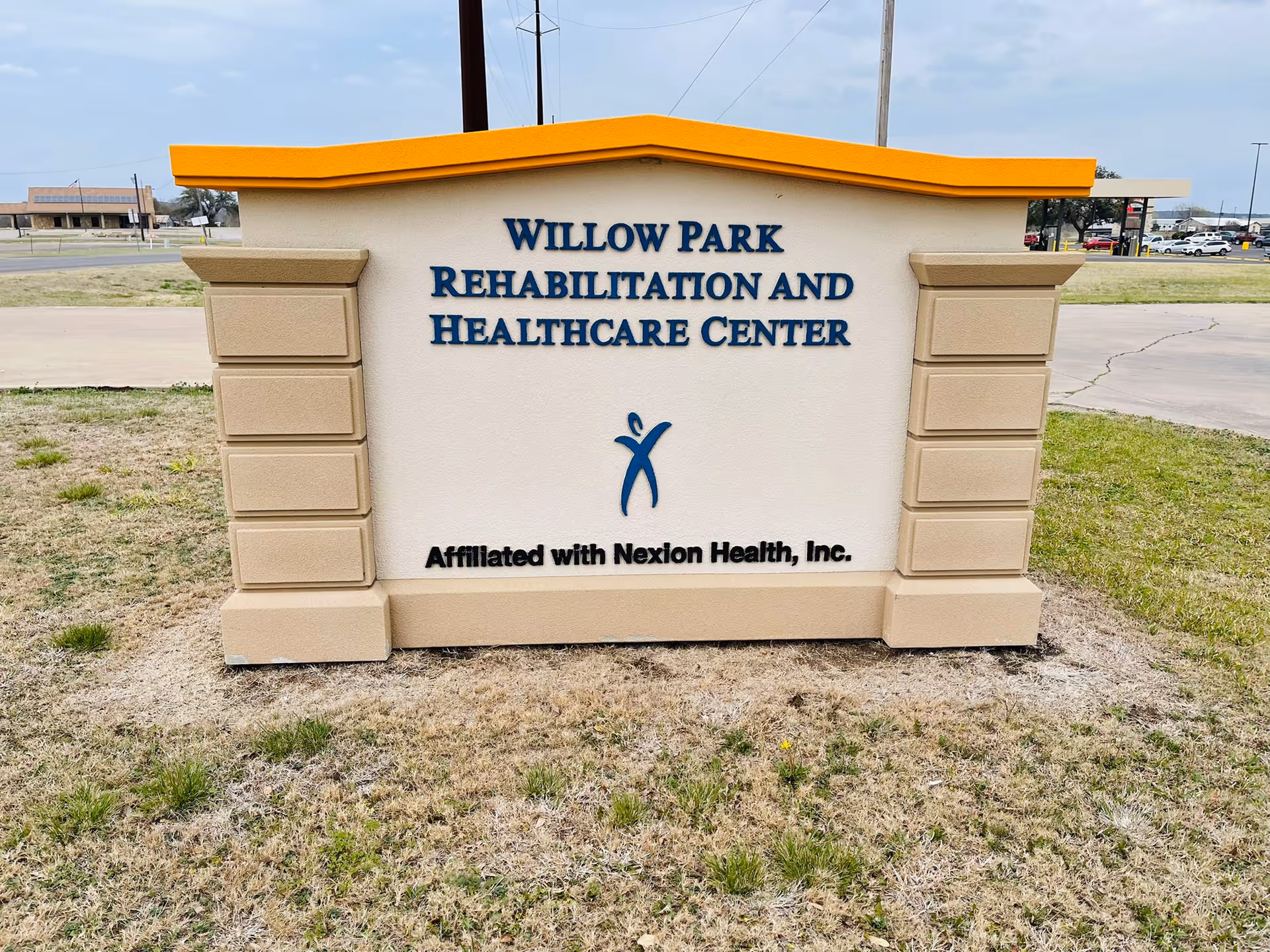 A large outdoor sign for Willow Park Rehabilitation and Healthcare Center with the text 'Affiliated with Nexion Health, Inc.' below the center name. The sign has a beige base with a yellow top and is situated on a grassy area near a paved road.