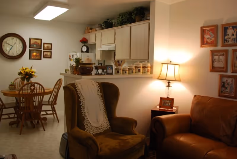 Cozy living room and dining area with upholstered chairs, a lit table lamp, framed photos, and a kitchen pass-through.