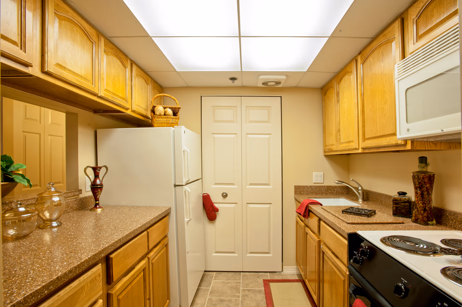 A compact kitchen with wooden cabinets, a white refrigerator, an electric stove with coil burners, a microwave, and a sink. The countertops are brown with a speckled pattern, and there are decorative items including a basket on top of the refrigerator, a vase, and glass jars on the counter. The kitchen has a tiled floor and a ceiling with fluorescent lighting panels.