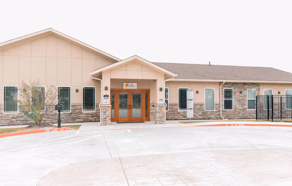 Exterior front view of BeeHive Homes of Rowlett building with beige walls, stone accents, multiple windows, and a double wooden door entrance under a small gabled roof.
