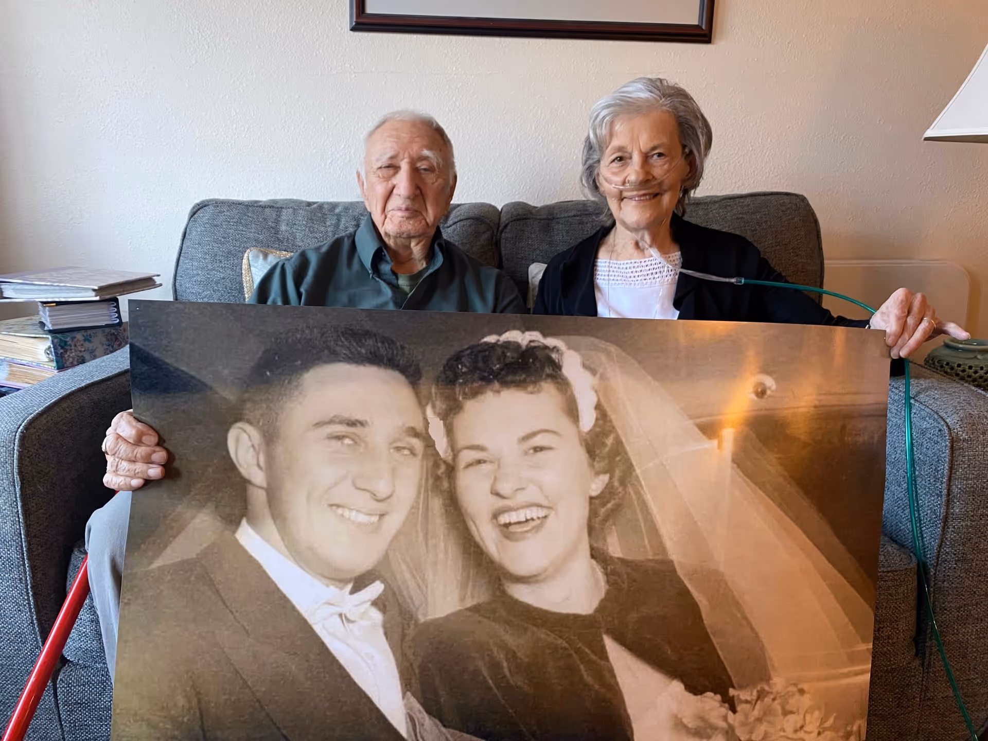 An elderly man and woman sitting on a gray couch holding a large black and white wedding photo of themselves. The woman is wearing an oxygen tube. There are books stacked on the armrest of the couch and a lamp on a side table.
