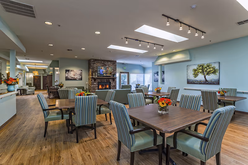 A spacious and well-lit dining area in a senior living facility with multiple wooden tables surrounded by striped upholstered chairs. The room features a brick fireplace with a TV above it, skylights in the ceiling, and several flower arrangements on the tables. The walls are painted light blue and decorated with paintings, including one of a tree. The floor is wooden, and there is a hallway visible in the background.