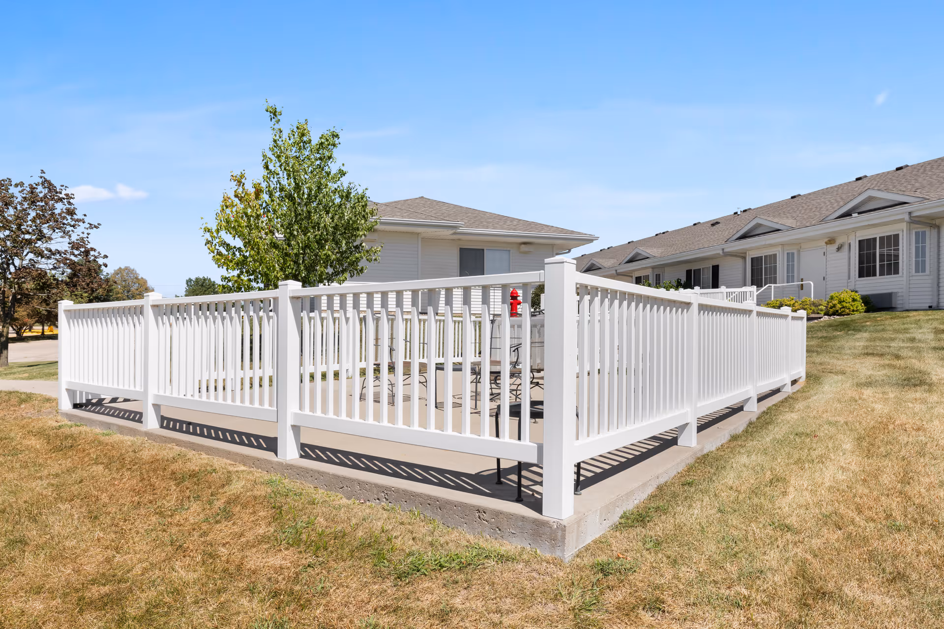 Outdoor patio area enclosed by a white picket fence with a few chairs and a table inside. The patio is part of a senior living facility with single-story buildings and a well-maintained lawn under a clear blue sky.