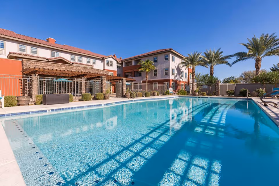 Outdoor swimming pool with clear blue water in front of a multi-story residential building with a red-tiled roof. Palm trees and a pergola with seating are visible around the pool under a clear blue sky.