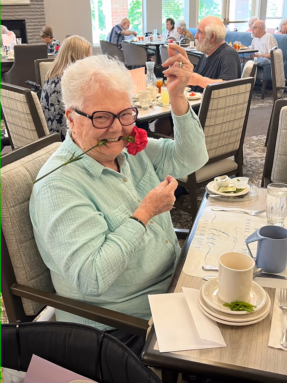 An elderly woman with white hair and glasses sits at a dining table in a senior living facility, holding a red carnation flower in her mouth and smiling playfully. Other elderly residents are seated at tables in the background, eating and conversing in a bright dining room with large windows.
