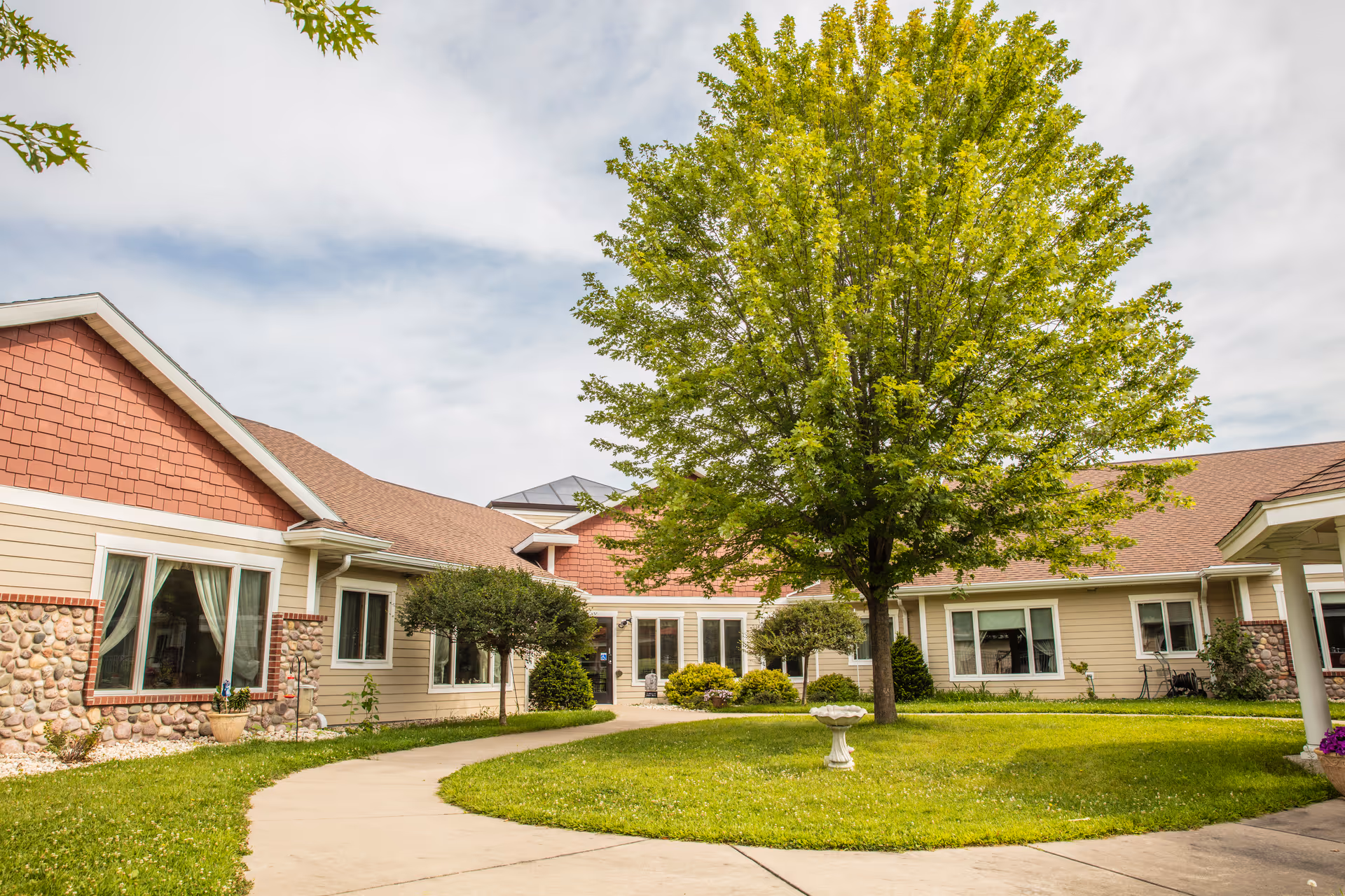Exterior view of Oakwood Manor senior living facility showing a courtyard with a large green tree in the center, surrounded by a curved concrete walkway and well-maintained grass. The building has beige siding with stone accents and red shingle roofing under a partly cloudy sky.