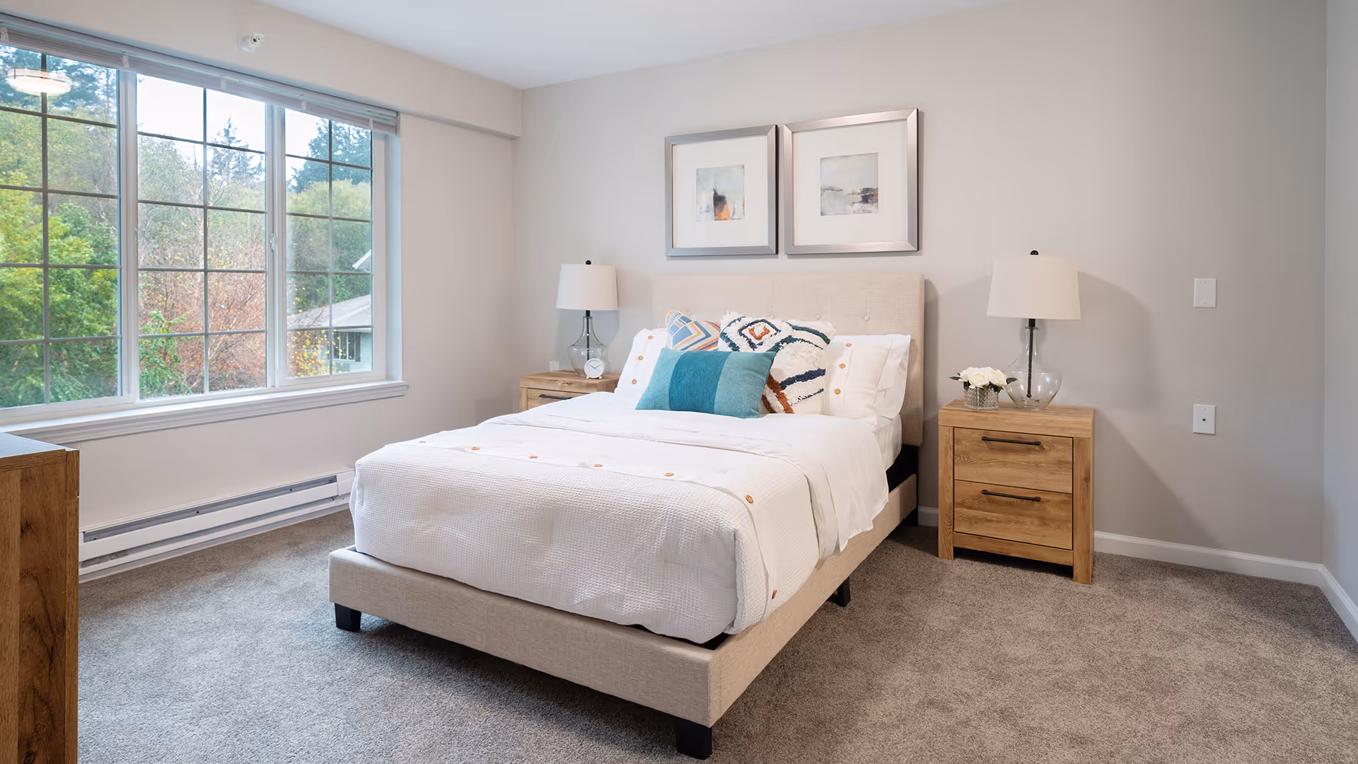 A bright furnished bedroom with a bed, bedside tables and lamps next to a large window overlooking trees.