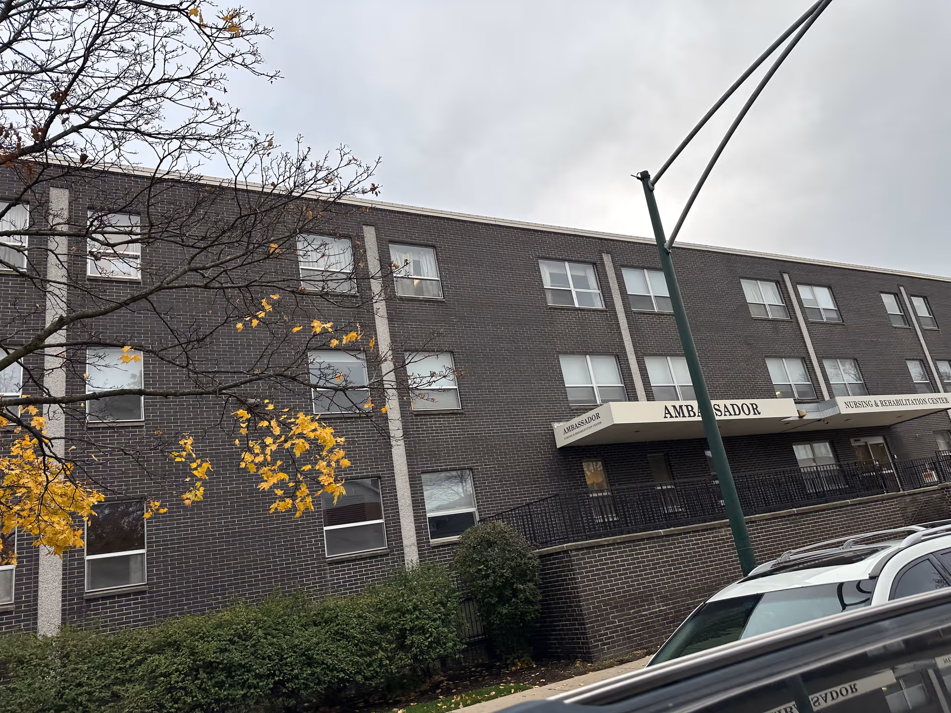 Exterior view of a three-story brick building with multiple windows, a tree with yellow leaves in the foreground, and a street lamp. The building has a sign that reads 'AMBASSADOR NURSING & REHABILITATION CENTER' above the entrance.