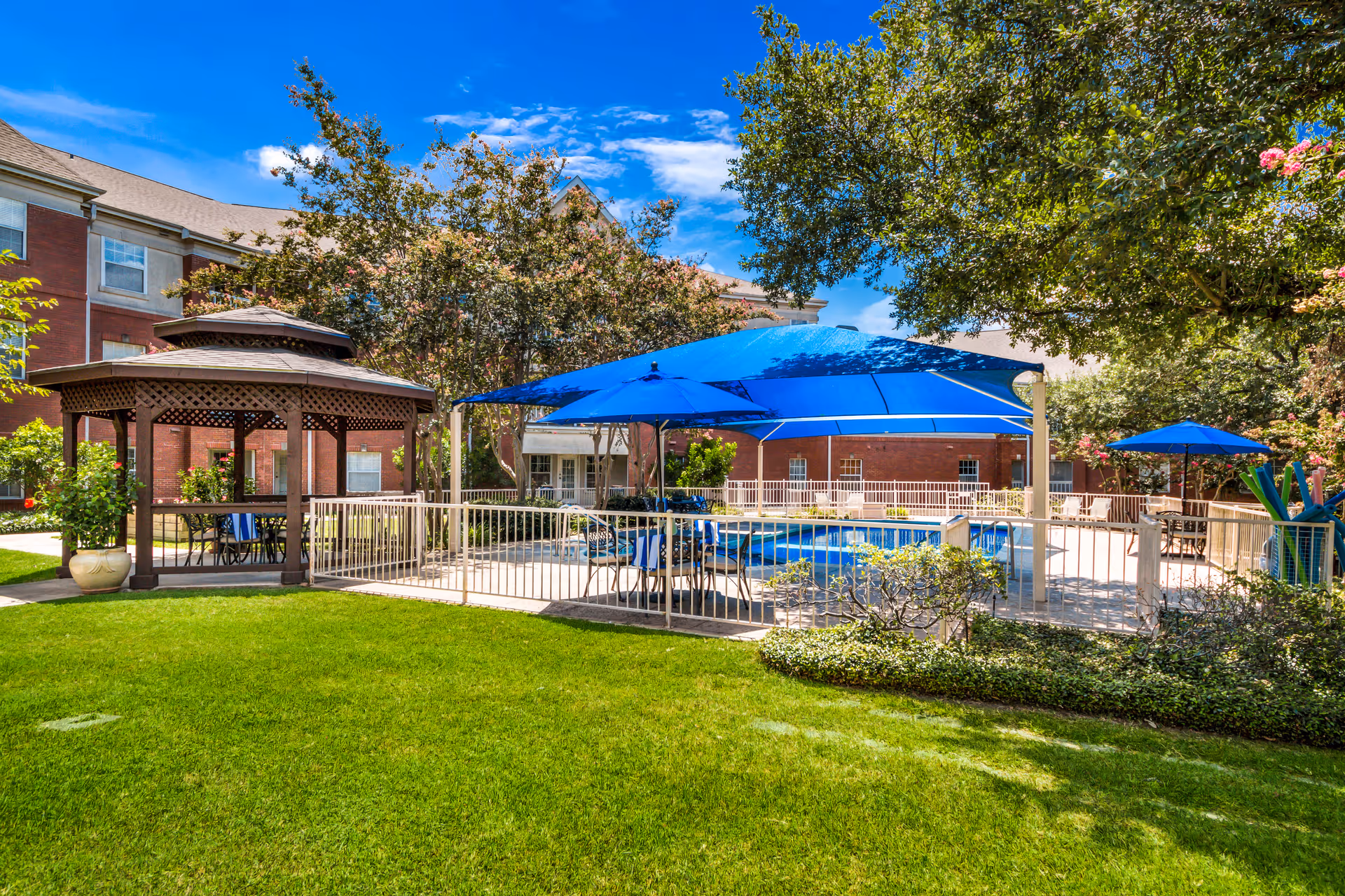 Outdoor area of The Forum at Lincoln Heights featuring a fenced swimming pool with blue umbrellas providing shade, a wooden gazebo with seating, lush green grass, trees, and a red brick building in the background under a bright blue sky.