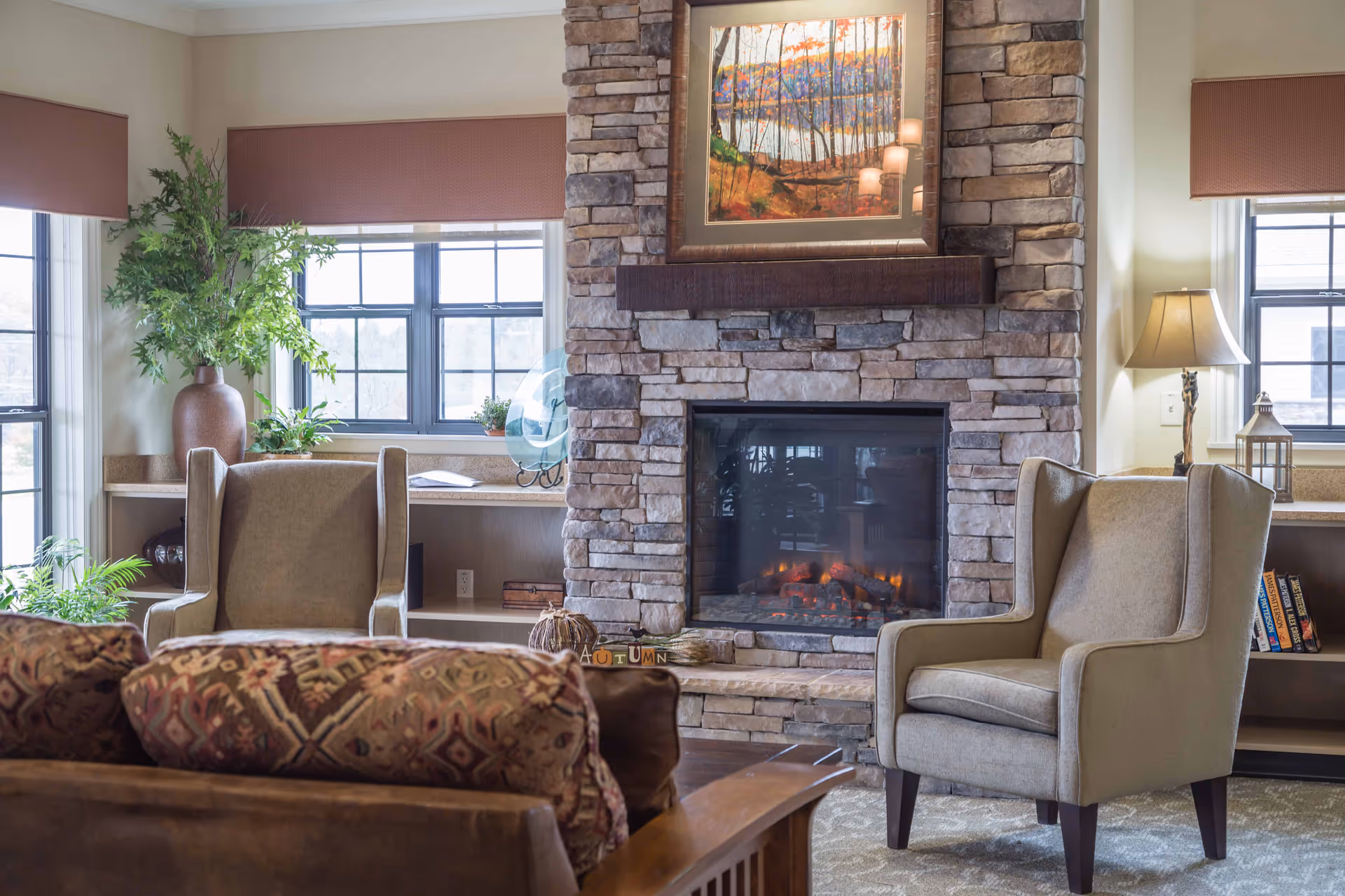 A cozy living room in a senior facility with a stone fireplace, two beige high-back chairs, a patterned sofa, plants, and large windows.