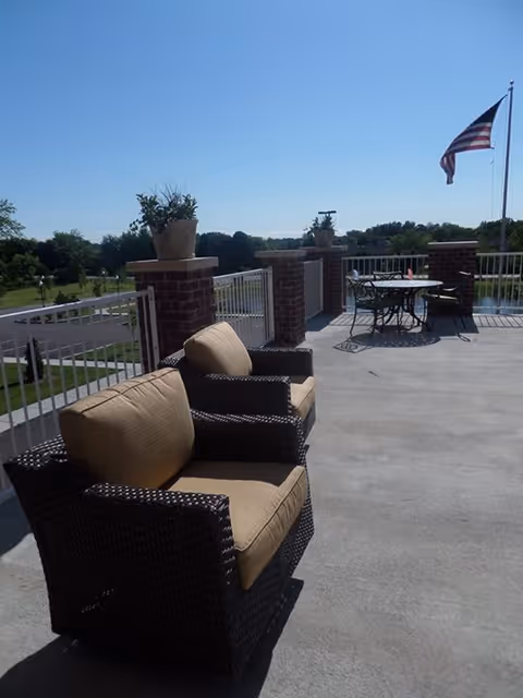 Sunny outdoor terrace with wicker cushioned chairs, a table by a railing, potted plants, and an American flag.