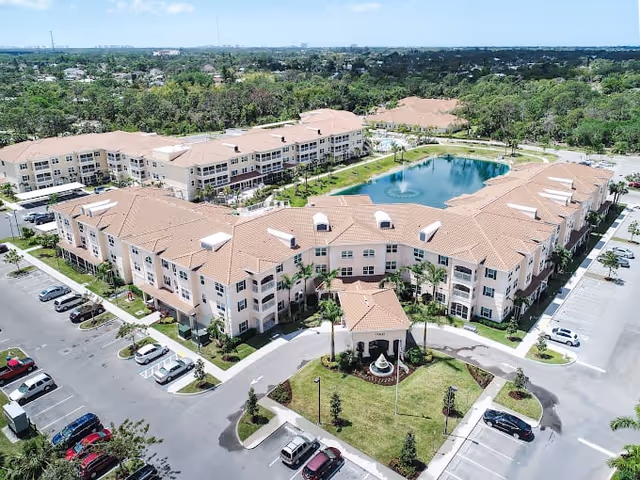 Aerial view of a large senior living facility with multiple connected buildings featuring beige walls and red-tiled roofs. The complex surrounds a pond with a fountain in the center. There are landscaped green areas, parking lots with cars, and a driveway leading to the main entrance with a covered drop-off area.