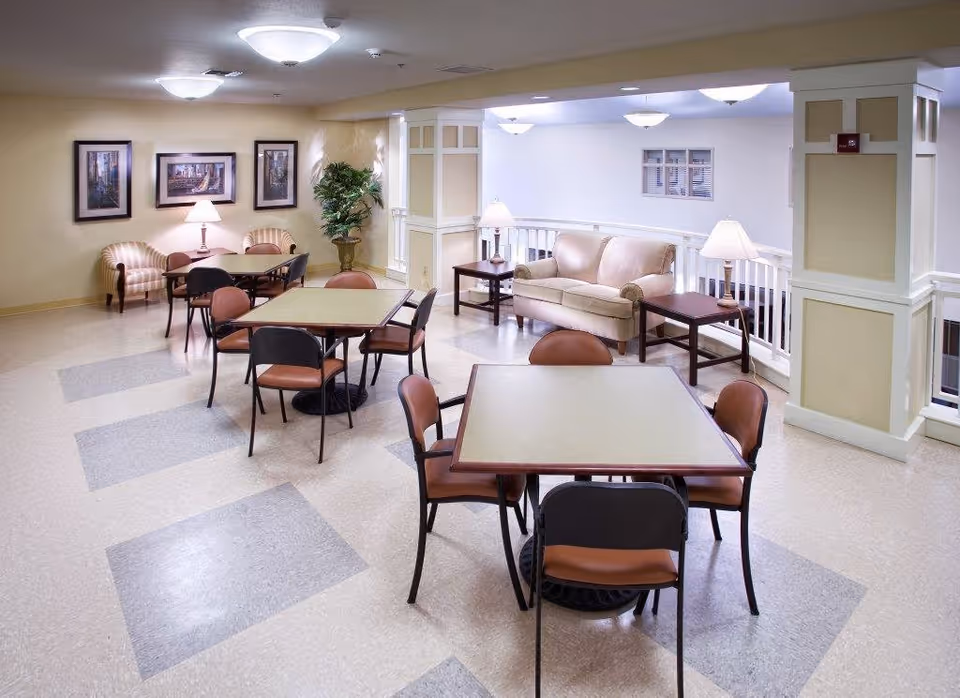 A well-lit common area in a senior living facility featuring several square tables with chairs arranged around them. There is a beige sofa with two side tables and lamps, a potted plant in the corner, and framed artwork on the wall. The floor has a pattern of light and darker tiles, and the space has a warm, inviting atmosphere.