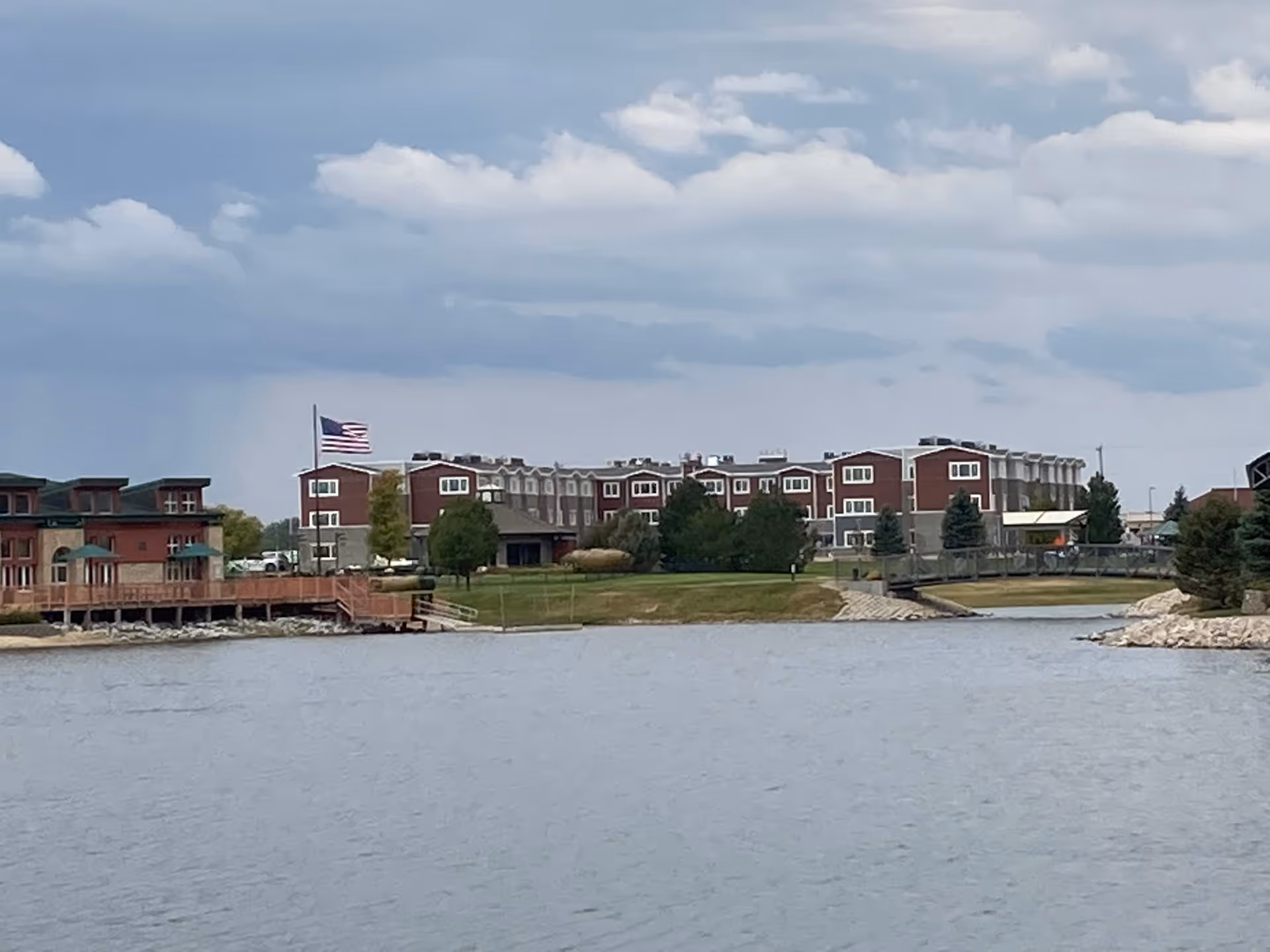 View of a senior living facility named Broadwell Senior Living from across a body of water, showing a large multi-story building with red and gray exterior, surrounded by trees and green lawn under a cloudy sky. An American flag is visible on a flagpole near the building.