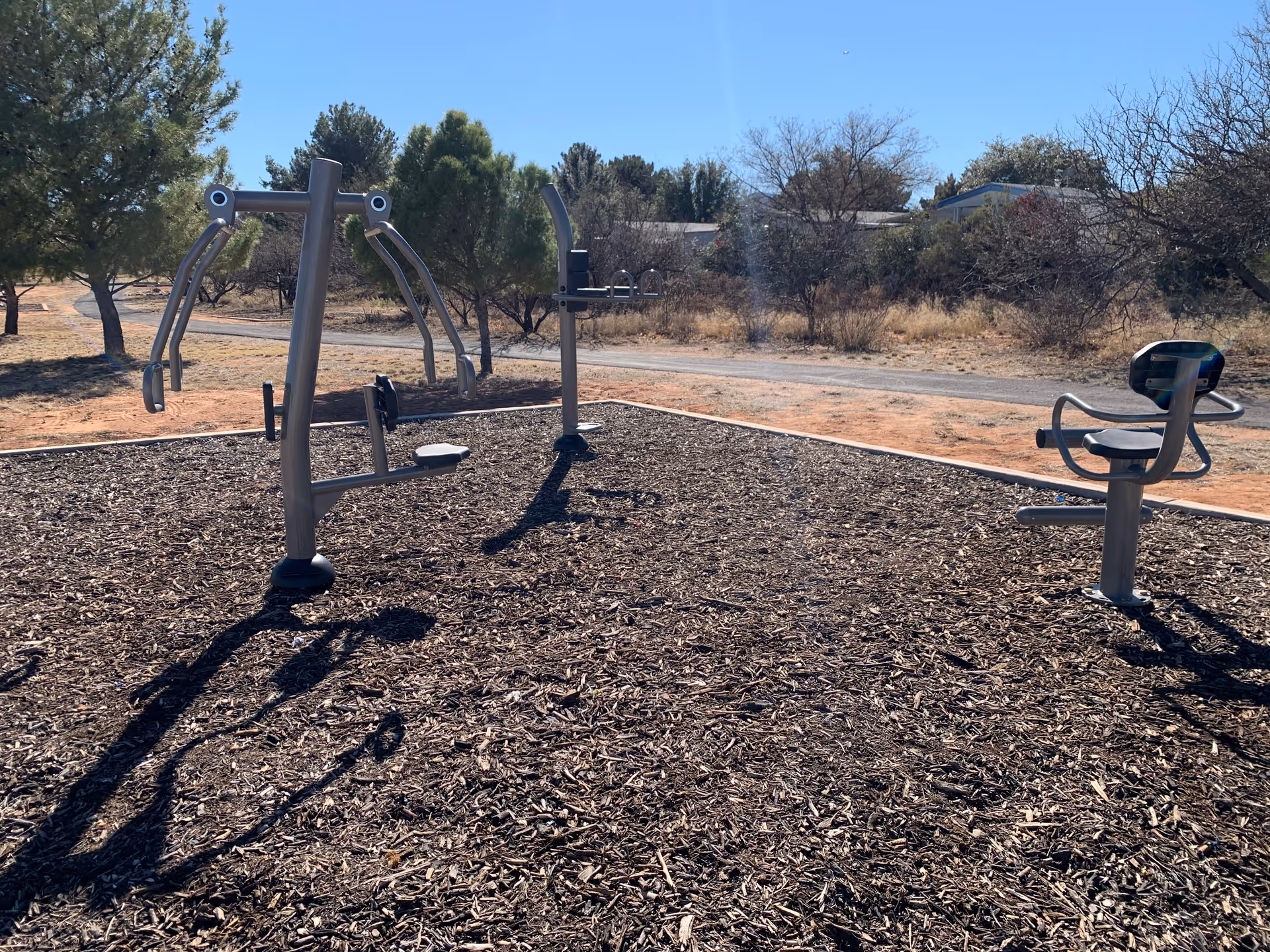 Outdoor fitness equipment set on wood chips with trees and a paved path in the background.