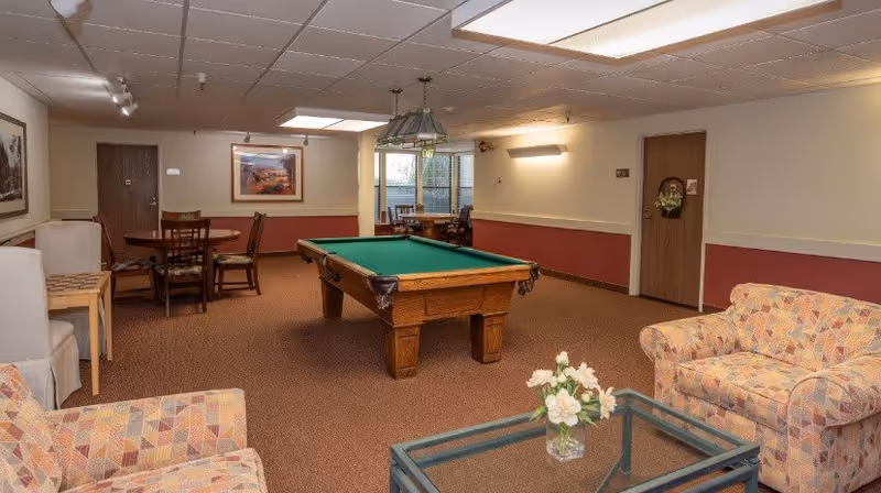 Communal lounge with a pool table in the center, patterned armchairs and a glass coffee table in the foreground, and a dining table in the background.