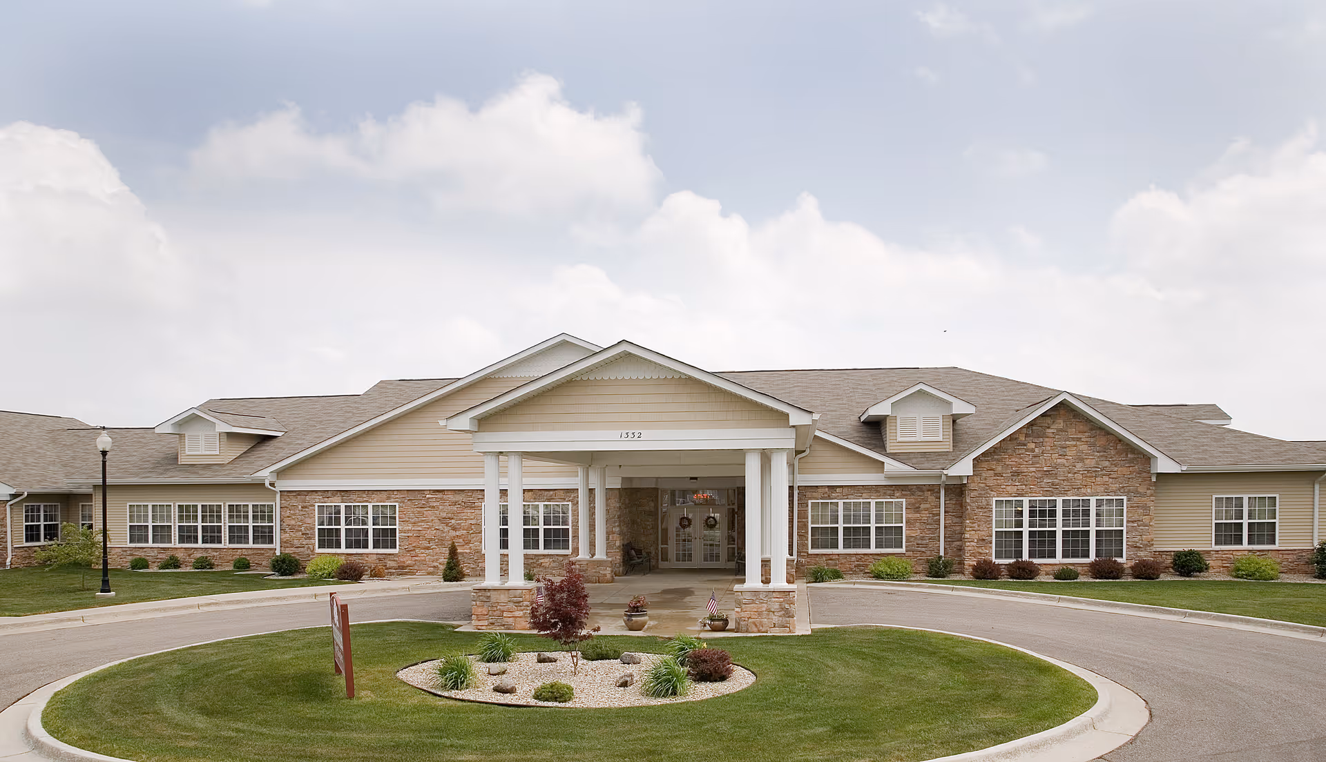Front exterior view of a single-story senior living facility building with a covered entrance supported by white columns, stone and beige siding, multiple windows, a circular driveway, and a landscaped area with grass and small plants under a partly cloudy sky.