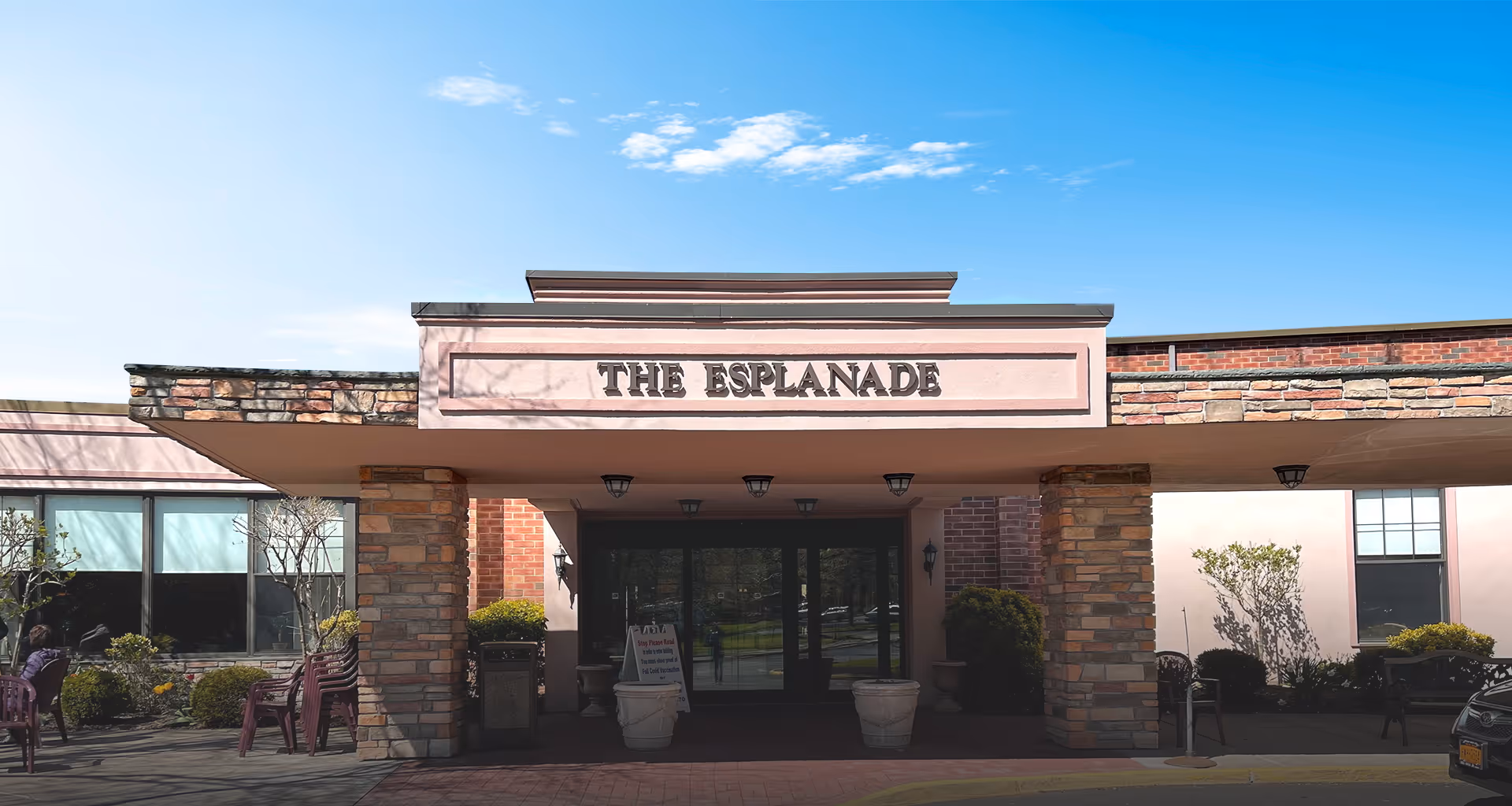 Front entrance of The Esplanade senior living facility with a covered drop-off area supported by stone pillars, outdoor seating with chairs and small bushes on either side, and a clear blue sky above.