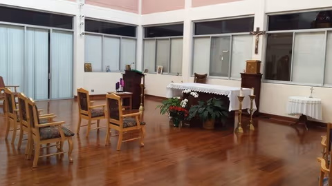 A small chapel or prayer room with wooden chairs arranged in rows facing an altar covered with a white cloth. The room has large windows with blinds, wooden flooring, and some plants near the altar. There are also candle holders and religious items on and around the altar.