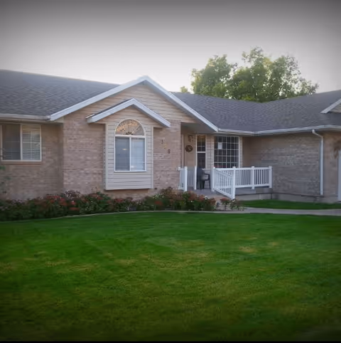 Exterior view of a single-story brick building with a well-maintained green lawn in front. The building has a small porch with white railings and a few chairs, a large window, and a smaller window with an arched top. Trees are visible in the background.