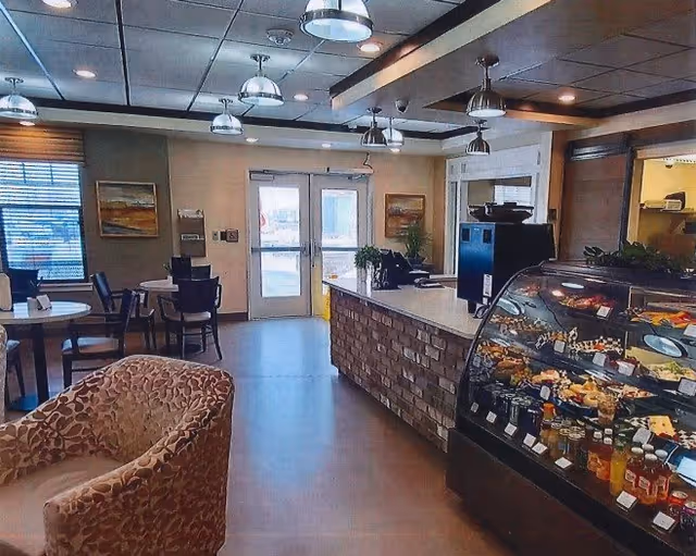 Interior view of a cozy dining area in a senior living facility with round tables and chairs, a leopard print armchair, a counter with a brick facade, and a glass display case filled with various food items. The room has pendant lights hanging from the ceiling and large windows letting in natural light.