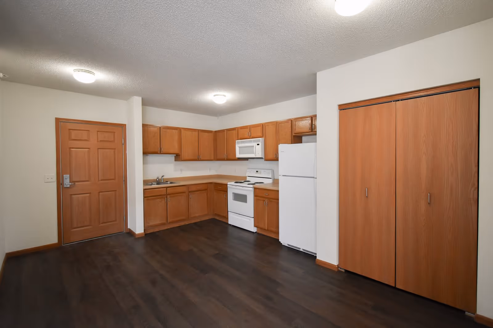 Interior view of a senior apartment kitchen area with wooden cabinets, a white refrigerator, stove, microwave, and a sink. There is a wooden door on the left and a double-door wooden closet on the right. The floor is dark wood and the walls are light-colored.