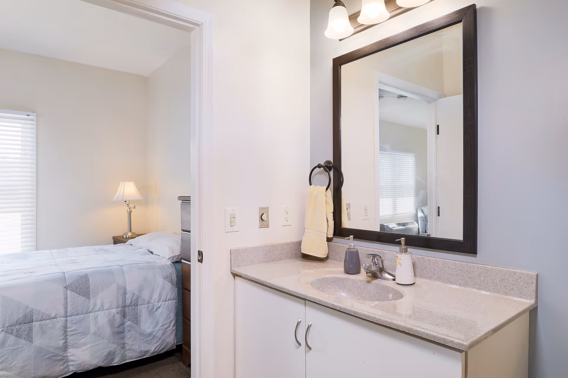 A view of a bedroom and bathroom vanity area in an assisted living facility. The bedroom has a bed with a geometric patterned comforter, a bedside table with a lamp, and a window with blinds. The bathroom vanity features a countertop with a sink, soap dispensers, a hand towel hanging on a ring, and a large mirror with three light fixtures above it.