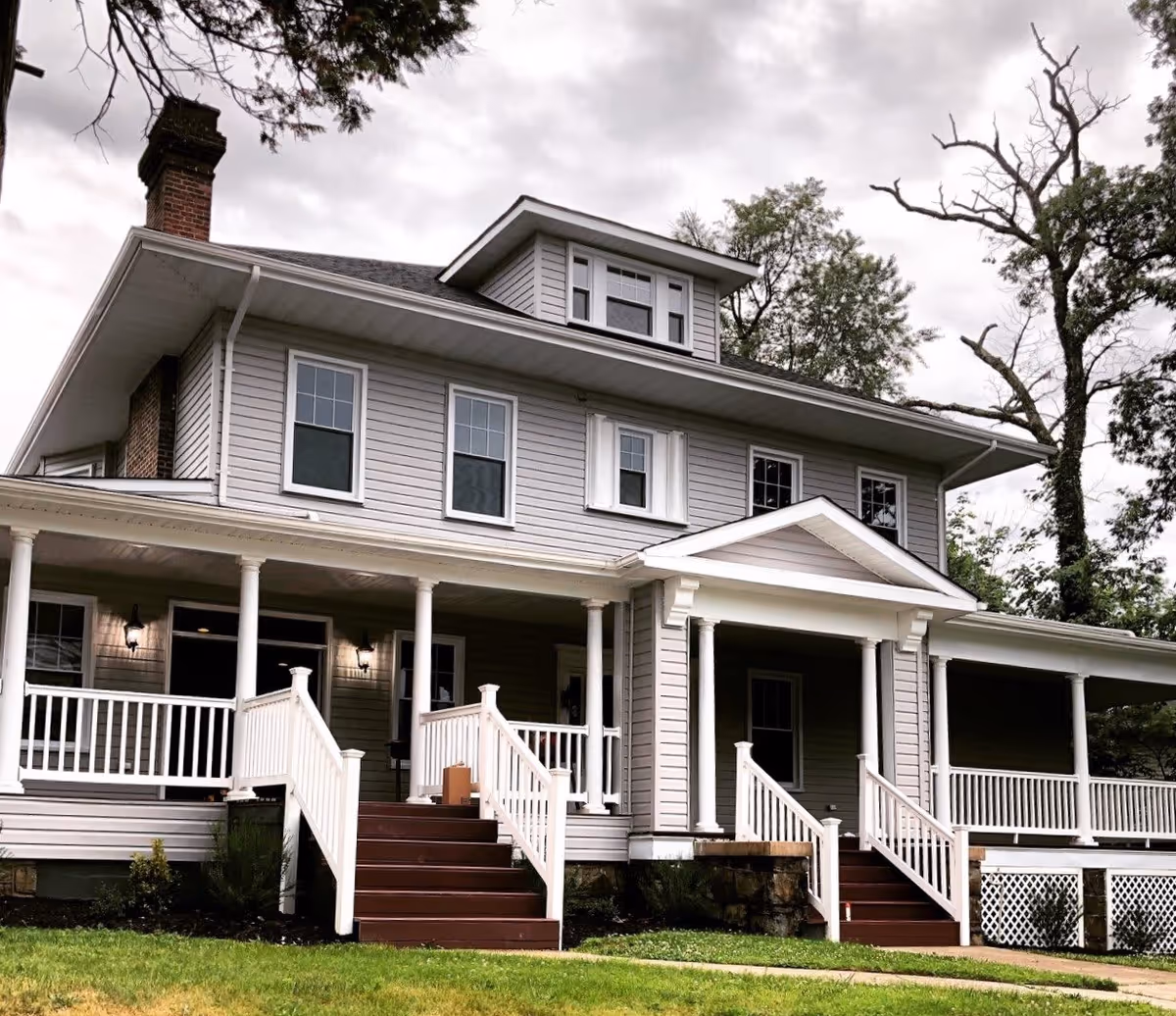 Exterior view of a two-story house with gray siding, white trim, and a large covered porch with white railings and steps leading up to the entrances. The house has multiple windows and a chimney, surrounded by trees and grass under a cloudy sky.