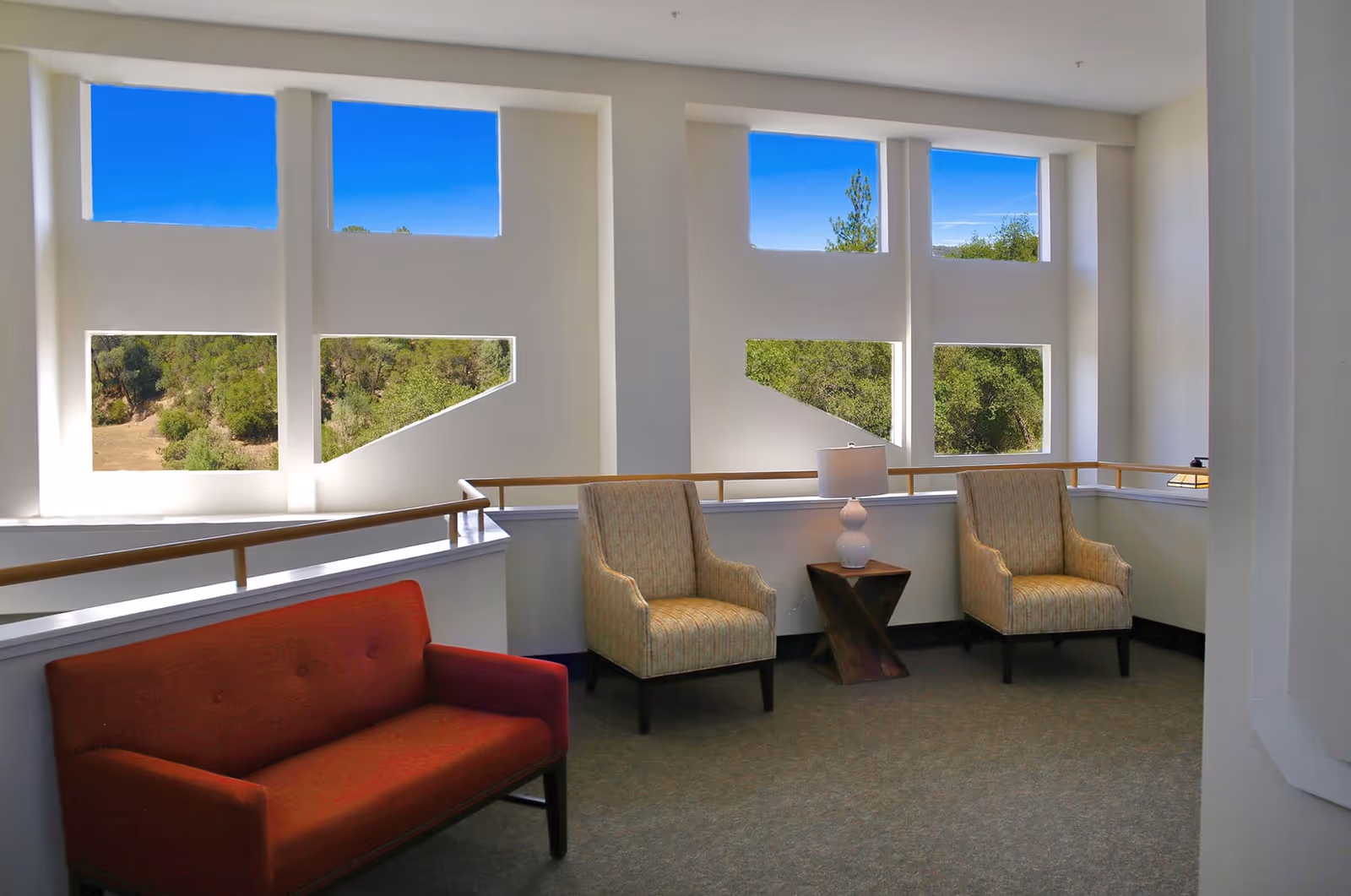 Sunlit seating area with a red loveseat, two upholstered armchairs, a side table and lamp in front of tall geometric windows overlooking trees.