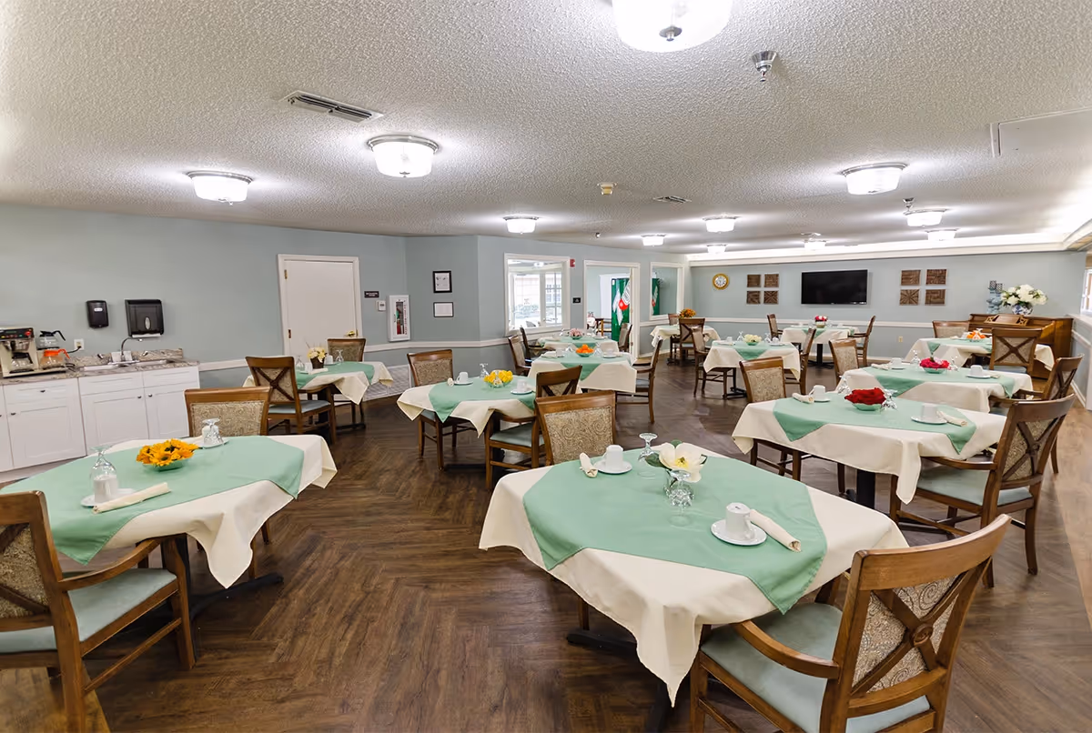 Spacious dining room with multiple tables set with green and white linens and wooden chairs, a serving counter along the wall and a TV at the far end.