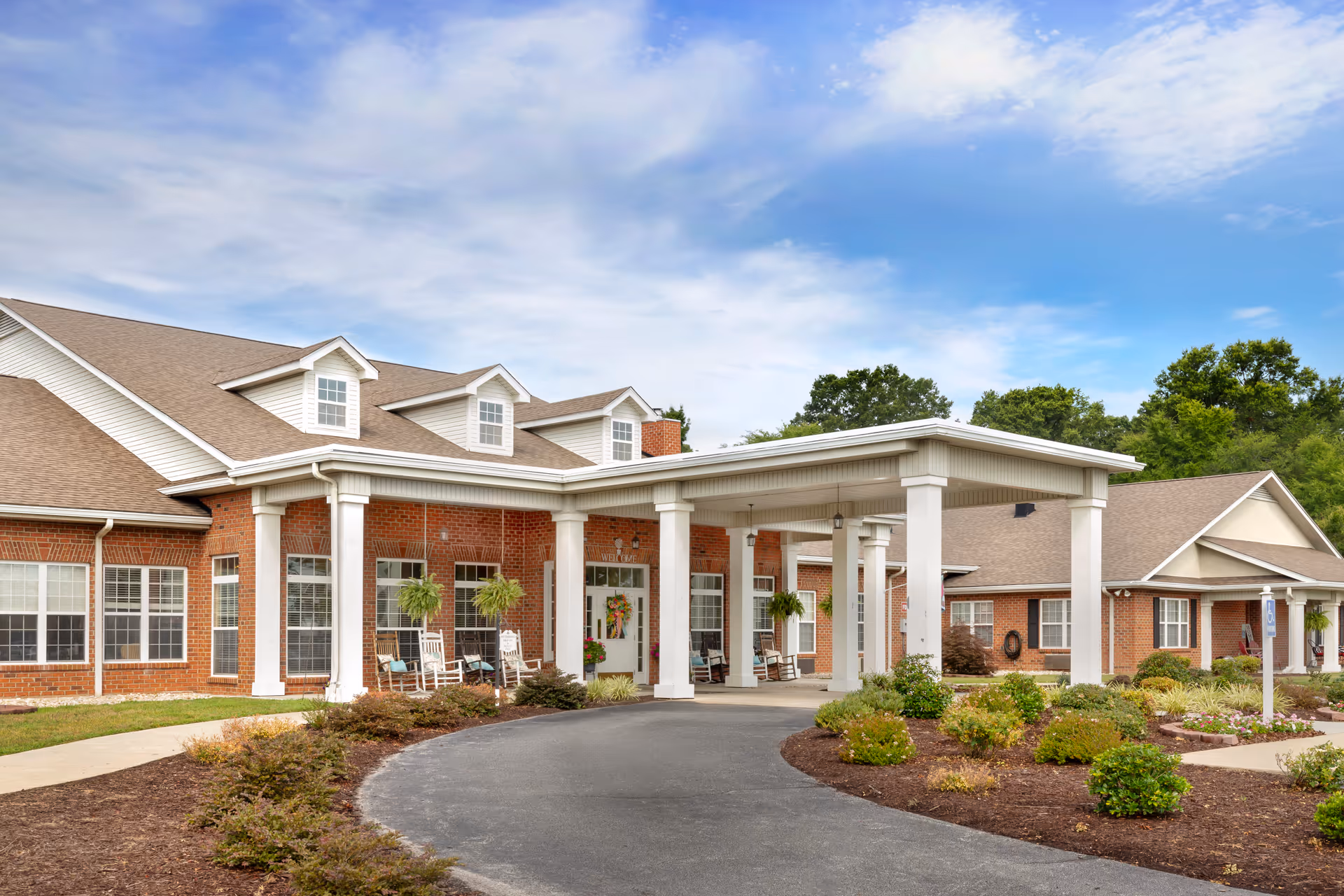 Front entrance of a brick senior living building featuring a covered porte-cochere with white columns, rocking chairs, and landscaped grounds.