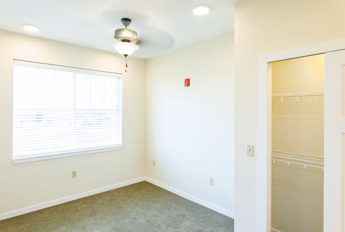 Empty room with beige walls, a large window with blinds, a ceiling fan with light, carpeted floor, and an open closet with wire shelving.