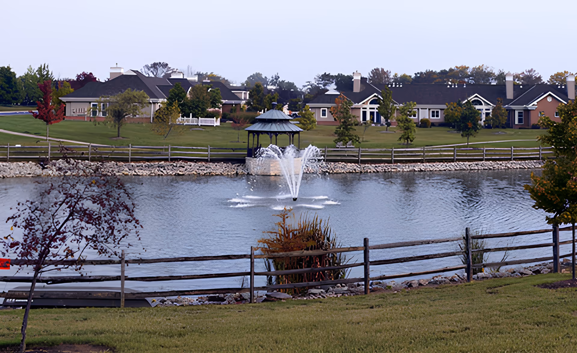 A serene outdoor scene at Knolls of Oxford featuring a pond with a central water fountain, surrounded by a wooden fence and landscaped greenery. In the background, there are residential buildings and a gazebo near the water's edge.