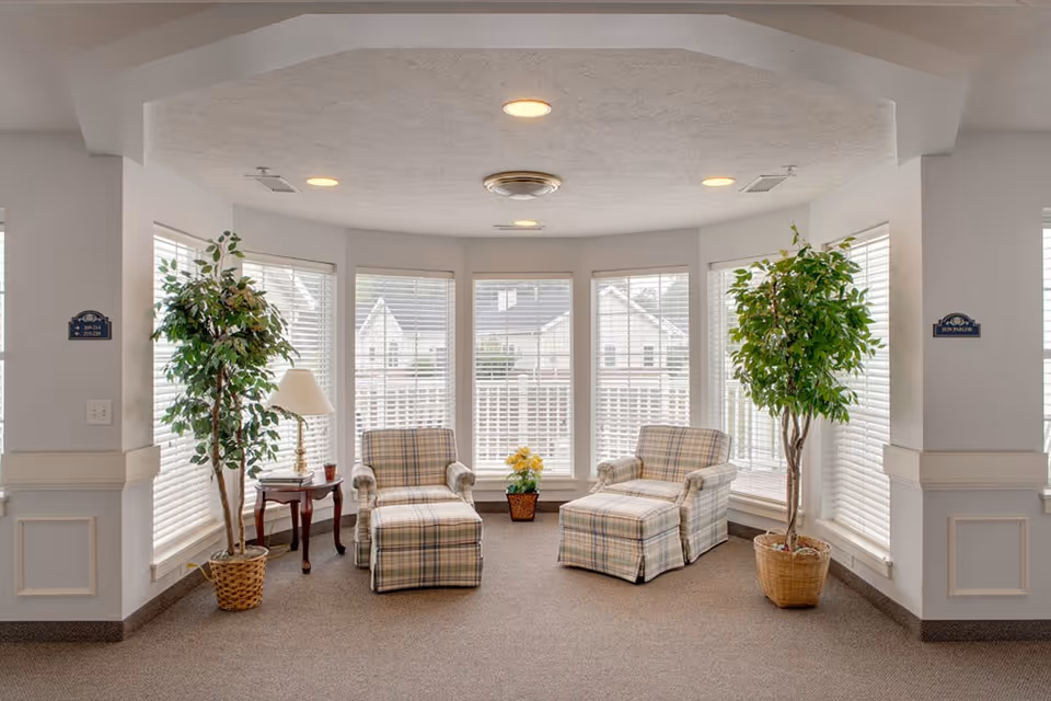 A bright sitting area with two plaid armchairs and matching ottomans positioned in front of large windows with white blinds. There are two tall potted plants on either side of the chairs, a small wooden side table with a lamp and a cup, and a small potted yellow flower in the center near the windows. The walls are white with decorative molding and there are ceiling lights above.