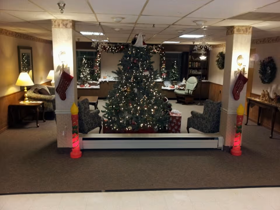 A cozy living room area decorated for Christmas with a large Christmas tree in the center adorned with lights and ornaments. Two red stockings hang on white pillars on either side of the tree, and two large candle decorations are placed at the base of each pillar. The room has comfortable chairs, a sofa, lamps, and festive wreaths on the walls. The lighting is warm and inviting.