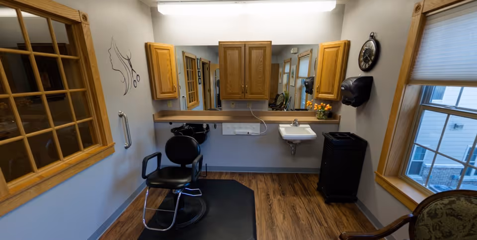 Interior view of a small hair salon area with a black salon chair on a black mat, a wall-mounted sink, wooden cabinets, a large mirror, a window with blinds, a wall clock, and a small vase of orange flowers on the counter.
