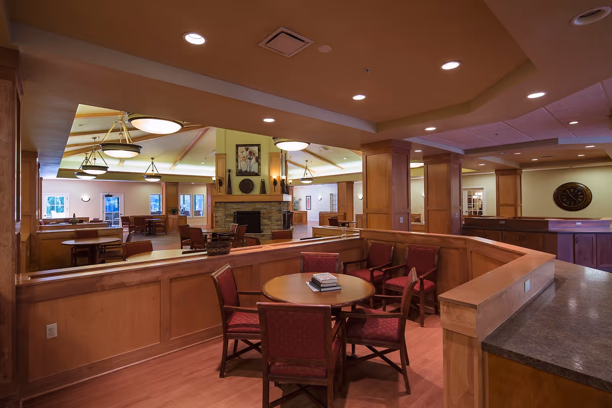 Interior view of a senior living facility common area with wooden furniture, round tables surrounded by red cushioned chairs, a stone fireplace with decorative items above it, and warm lighting from ceiling fixtures.