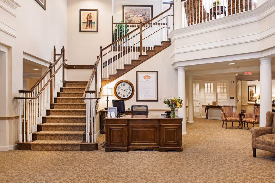 Lobby with a wooden reception desk, carpeted double staircase, and seating area in a senior living facility.
