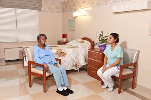 An elderly woman sitting in a chair smiling and talking with a healthcare worker who is seated in another chair in a room with a bed, nightstand, and window with blinds.