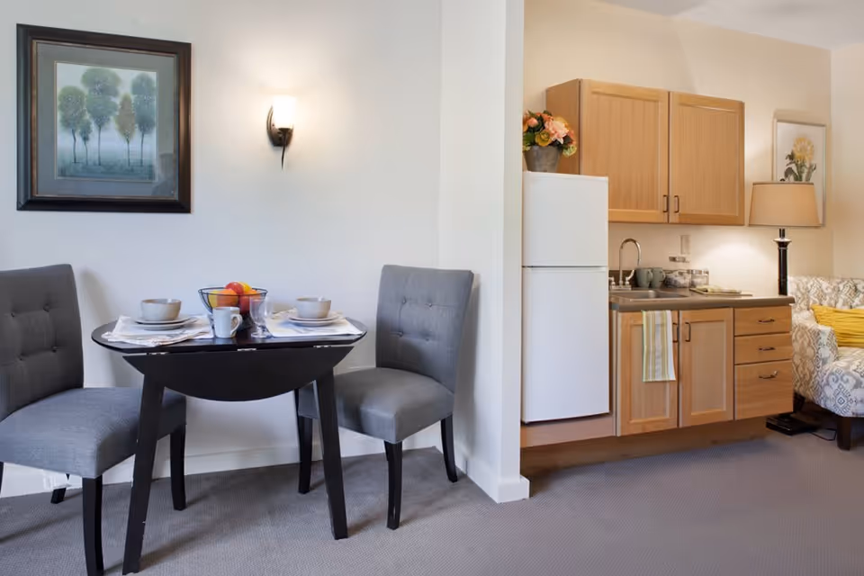 A small dining area with a black table set for two, featuring gray upholstered chairs, bowls, cups, and a fruit bowl. Adjacent to the dining area is a kitchenette with wooden cabinets, a white refrigerator, a sink, and a countertop with a lamp and decorative flowers. A framed painting and a wall sconce light are visible on the wall.