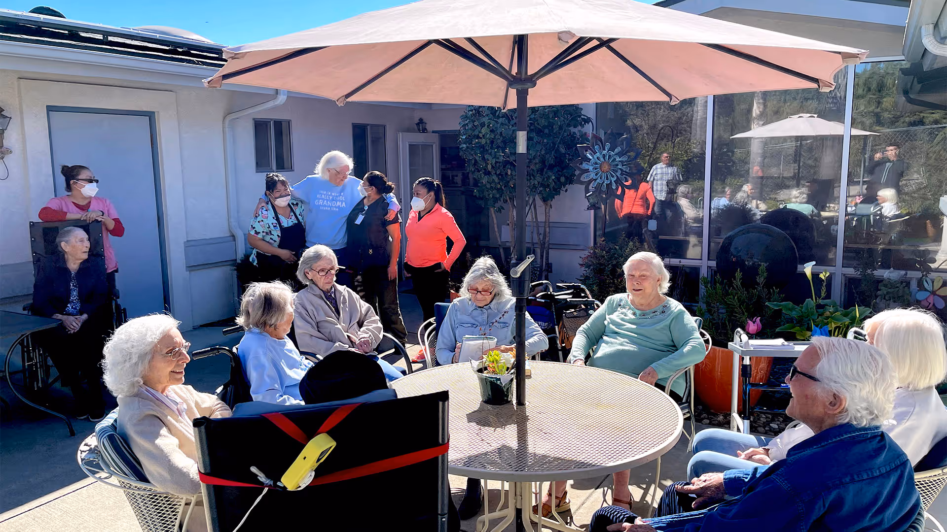 Elderly residents and caregivers gathered outdoors around a round patio table under a large umbrella on a sunny day.