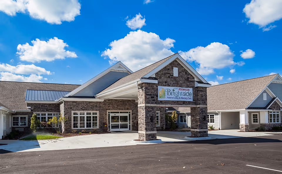 Exterior front view of Brightside Assisted Living & Memory Care facility with a covered entrance supported by stone pillars, a clear blue sky with scattered clouds in the background, and a paved driveway in front.