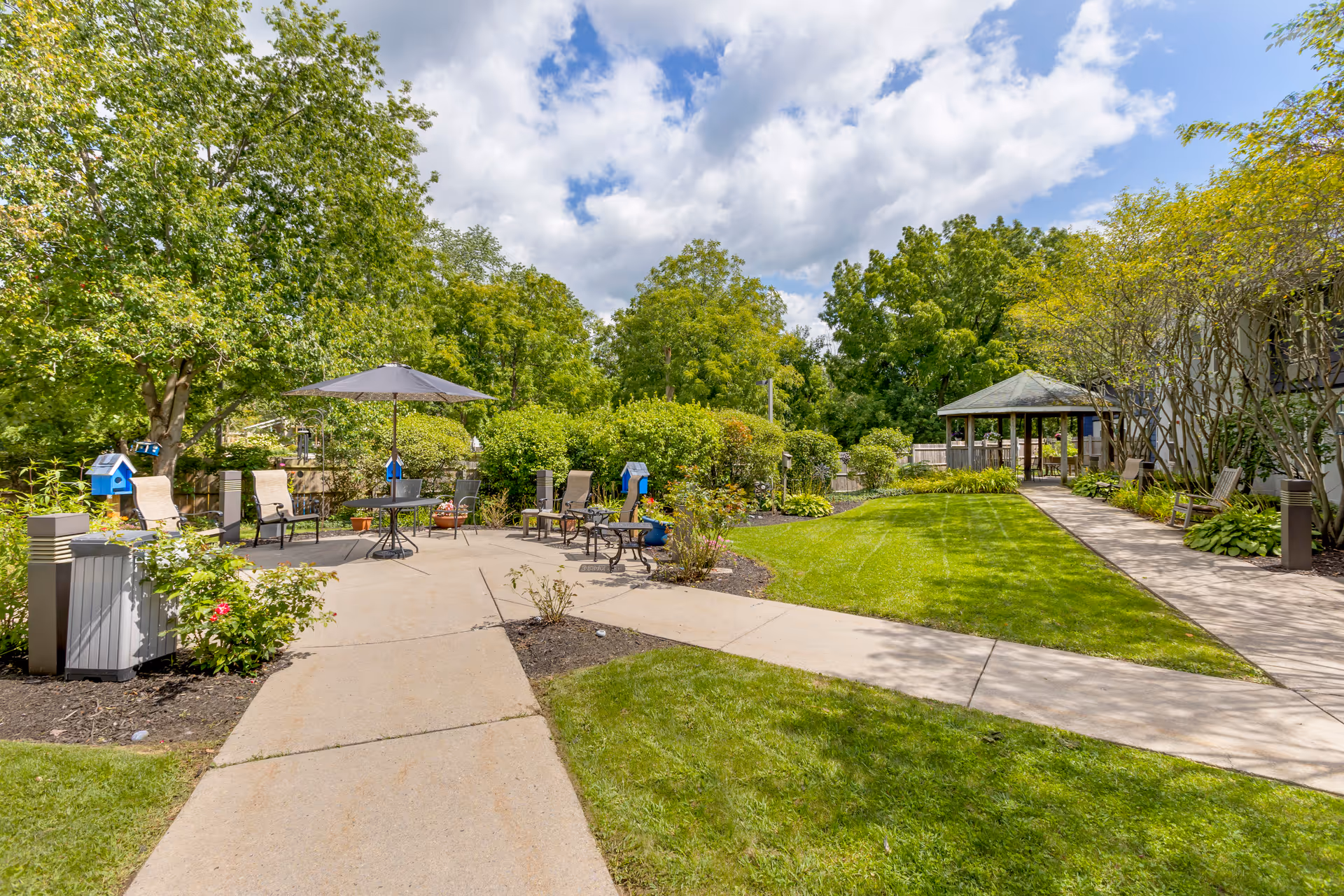 Outdoor patio area with several chairs and a table under a large umbrella, surrounded by green trees and bushes. There is a paved walkway leading to a gazebo on the right side, with well-maintained grass and garden beds along the paths.