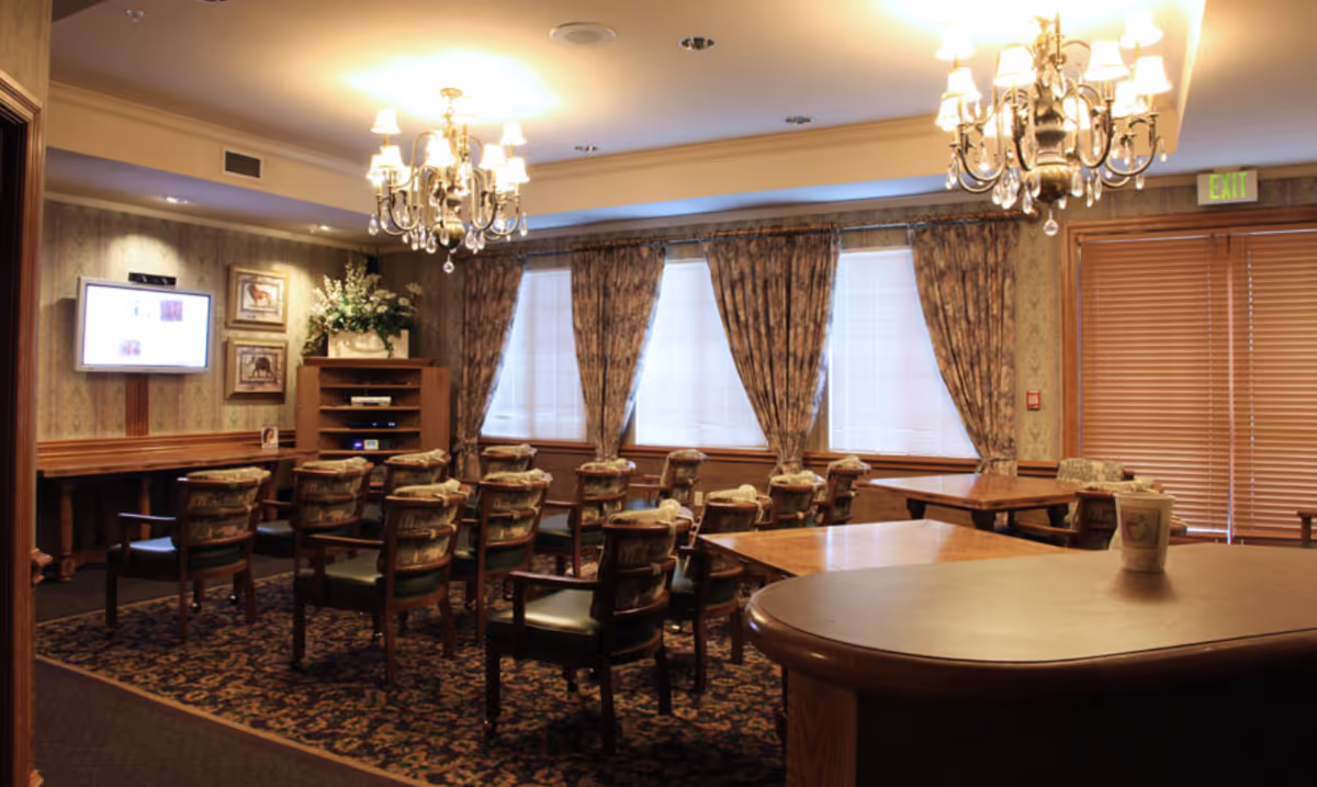 Well-lit communal dining/activity room with chandeliers, rows of chairs and tables facing a wall-mounted TV and draped windows.