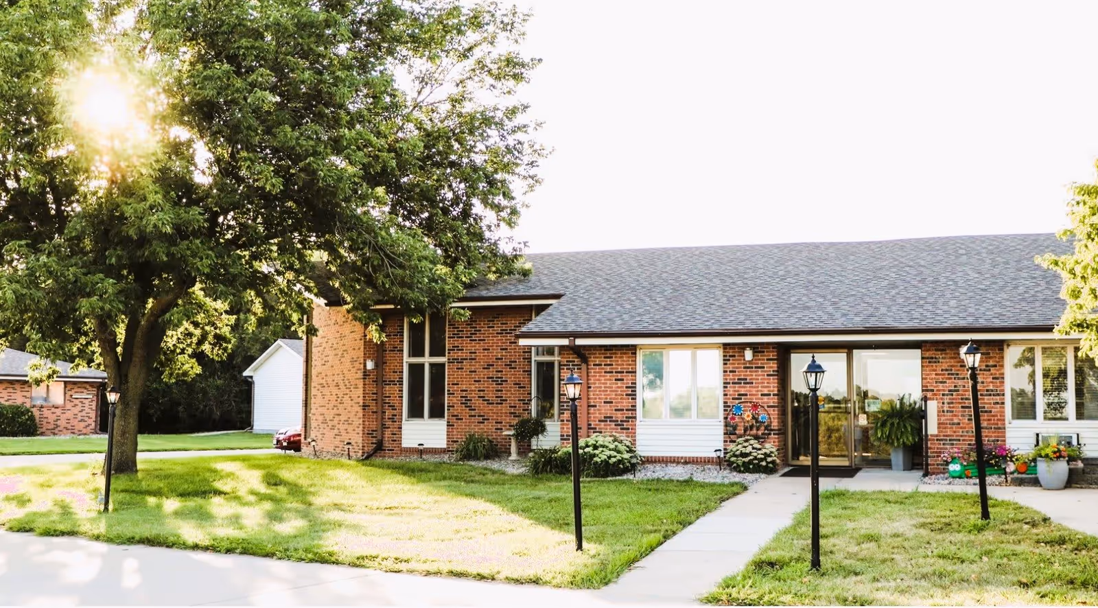 Exterior view of a single-story brick building with a gray shingled roof, surrounded by green grass and trees. A concrete pathway leads to a glass entrance door flanked by black lamp posts. There are flower pots and decorative plants near the entrance.